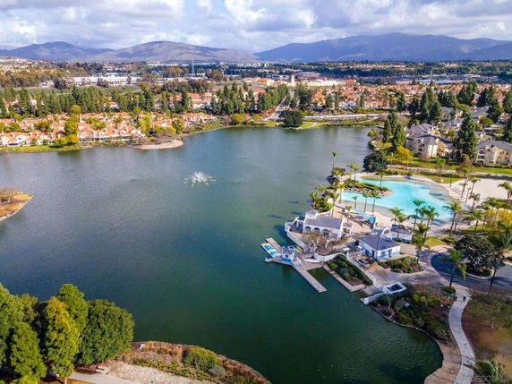 Panoramic aerial view of a lake surrounded by houses and trees, with hills in the background.