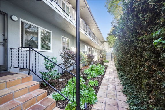 View of a pathway beside a multi-story apartment building with plants and bushes.
