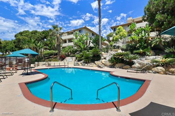 Outdoor swimming pool surrounded by lounge chairs and umbrellas, with apartment buildings in the background.