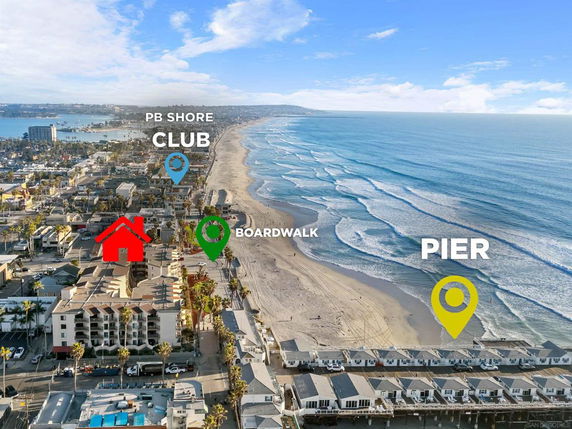 Aerial panoramic view of a beachfront area with buildings, a boardwalk, and a pier by the ocean.