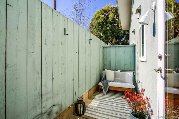 Narrow outdoor space with a wooden bench, striped rug, and decorative plants next to a building.