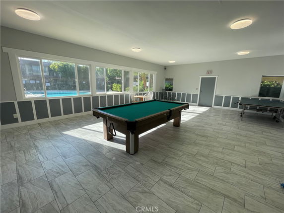Room with a billiards table and tile flooring, featuring large windows providing a view of an outdoor pool area.