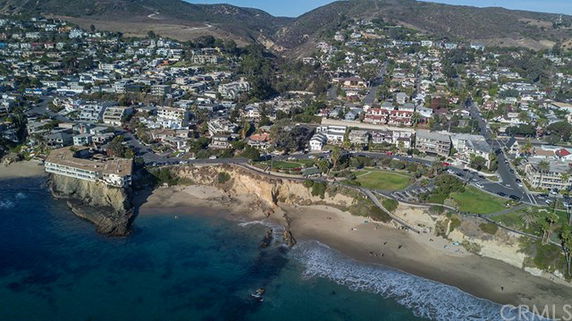 Aerial panoramic view of a coastal town with buildings on a hillside overlooking a beach and ocean.
