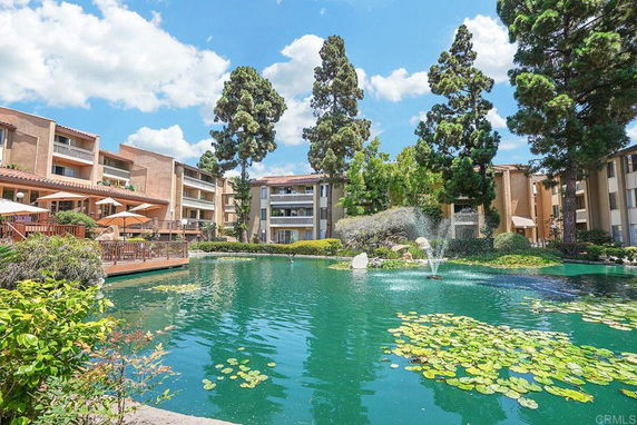 Exterior view of multi-story apartment buildings surrounding a landscaped pond with fountains and trees.