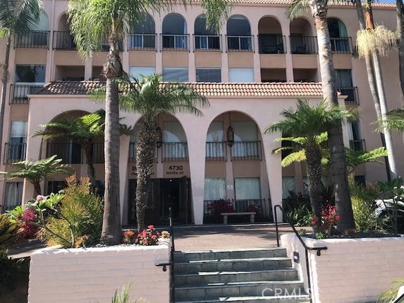 Front view of a multi-story building with arches and balconies, surrounded by palm trees and landscaped plants.
