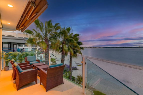 Outdoor seating area on a balcony overlooking a beachfront with palm trees and a view of the water at sunset.
