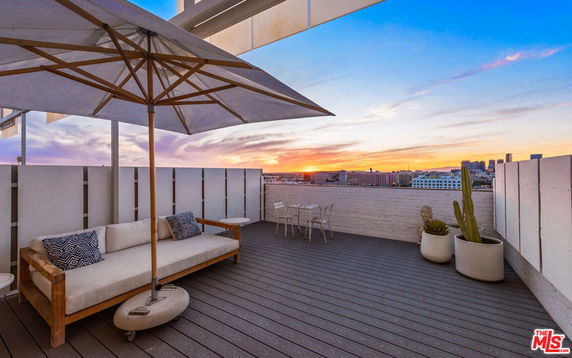 Rooftop with lounge seating, umbrella, and plants overlooking a cityscape at sunset.