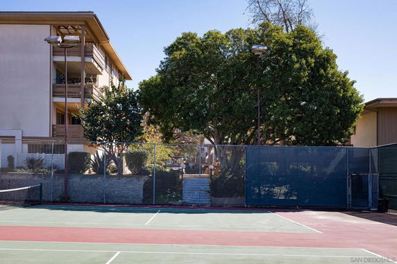 Tennis court adjacent to a multi-story building and surrounded by trees.
