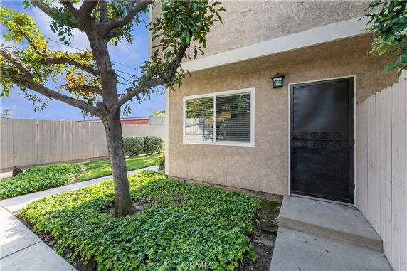 Front view of a house with a single door and window, surrounded by greenery and a fence.