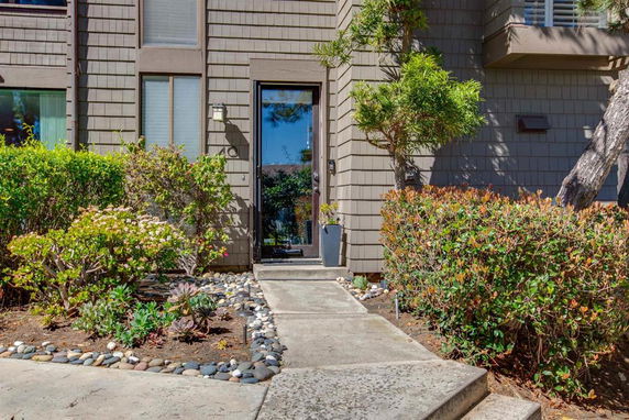 Front view of a residential building with shingle siding and a glass entry door, surrounded by bushes and landscaped plants.