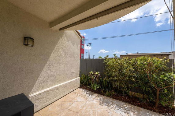 View from a shaded outdoor patio area with a fence and some plants visible beyond it.
