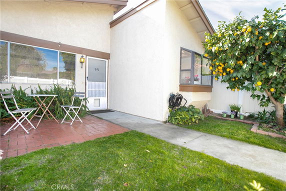 Front view of a house with a small patio, a door, and a window. There is a table and chairs on the patio, and a lemon tree on the side.