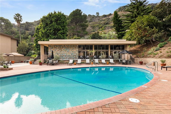 Outdoor swimming pool with poolside chairs and tables, surrounded by trees and a building in the background.