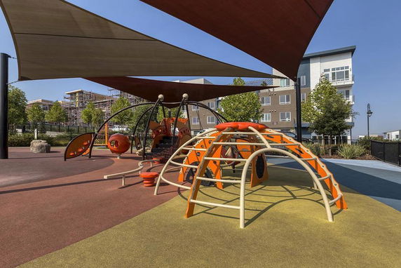Outdoor playground with climbing structures and shade, adjacent to residential buildings.