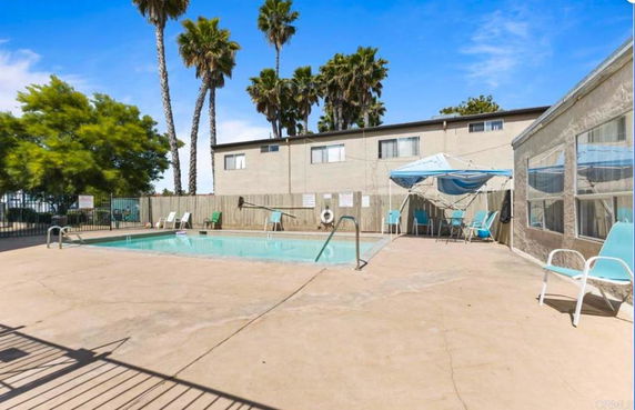Outdoor swimming pool area with lounge chairs and a sunshade canopy beside a two-story building.