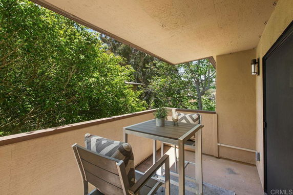 Covered outdoor balcony with a table and chairs, overlooking lush green trees.