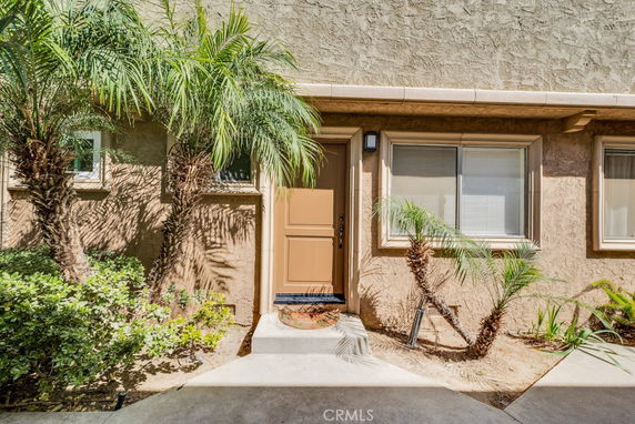 Front view of a house with a single door and windows, surrounded by small palm trees.