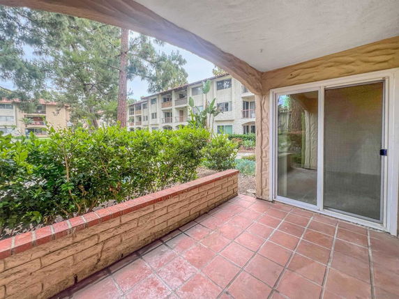 View from a covered patio area looking out onto a garden with shrubs and trees in front of a multi-story residential building.