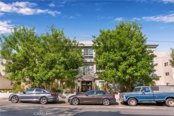 Front view of a multi-story residential building with parked cars and trees in front.