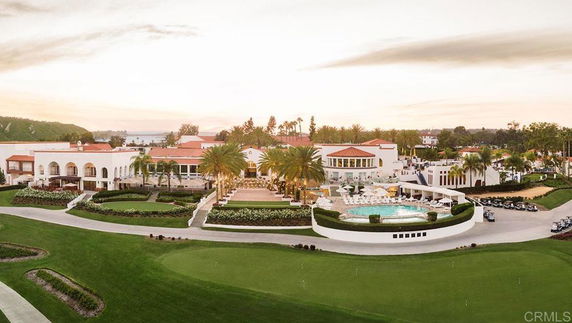 Aerial view of a large resort complex with multiple white buildings, a round swimming pool, palm trees, and landscaped gardens.