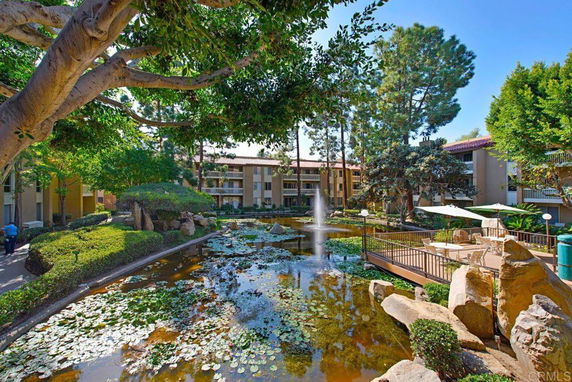 View of an apartment complex with a central water feature surrounded by trees and greenery, showing multiple apartment buildings in the background.
