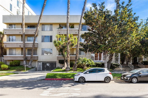 Front view of a multi-story apartment building with balconies and large trees in front.