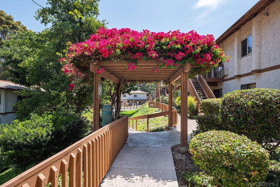 Pathway leading through a garden area with a flowering pergola and adjacent buildings.