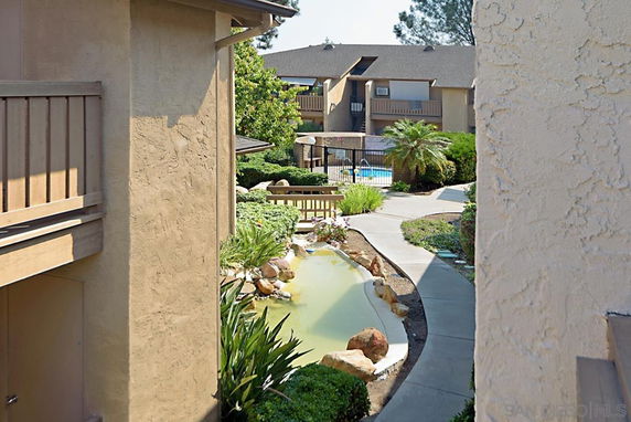 Outdoor area with a small pond, pathway, and a view of buildings in the background leading to a swimming pool.