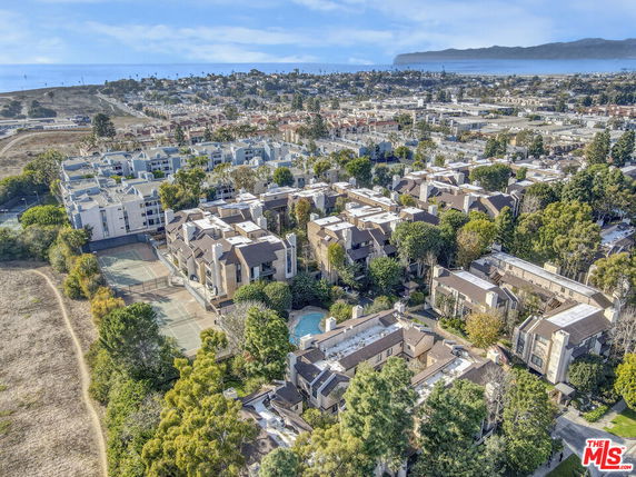 Aerial view of a residential area with multiple apartment buildings, surrounded by greenery, and a distant view of a cityscape with a coastline.