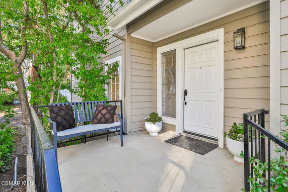 Front view of a house entrance with a white door and a glass panel, featuring a small porch with a bench and potted plants.