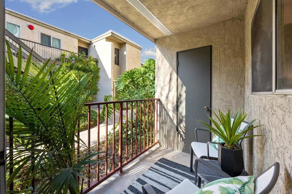 View from a covered balcony with potted plants and chairs, overlooking an apartment complex.
