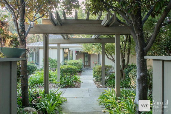 Front view of a house with a wooden pergola entrance and surrounding shrubbery.