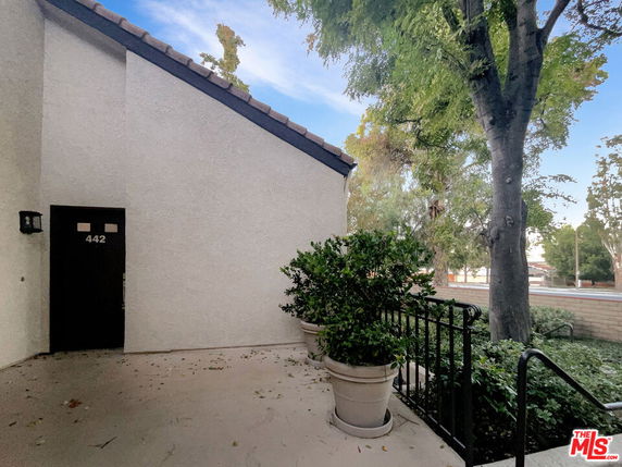 View of a building entrance with a sloped roof and a black door, surrounded by plants and trees.