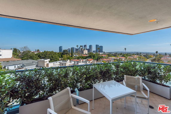 View of a cityscape from a balcony with chairs and a table, overlooking buildings and greenery.