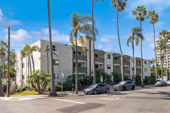 Front view of a multi-story apartment building with balconies and palm trees along the street.
