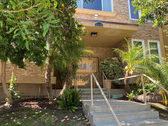 Front view of a house with stairs leading to the entrance, surrounded by plants and trees.