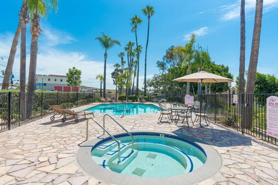 Outdoor area with a swimming pool and a hot tub on a stone patio, surrounded by palm trees and fenced in with lounge chairs and tables.