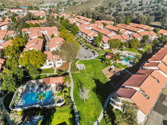 Aerial view of a residential complex with red-roofed buildings, landscaped green spaces, swimming pools, and surrounding hilly terrain.