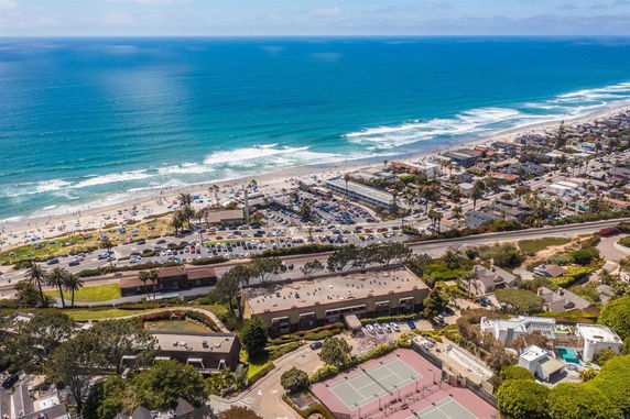 Aerial view of a coastal area with a beach, parking lot, and nearby buildings.