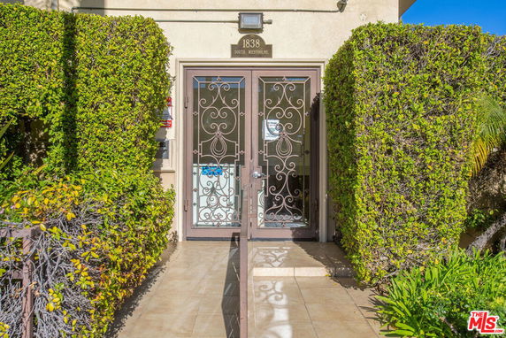 Front view of a building entrance with ornate metal doors and trimmed hedges.