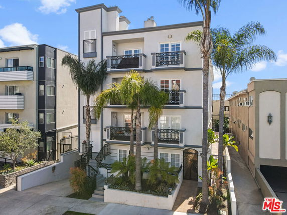 Front view of a three-story residential building with balconies and palm trees.