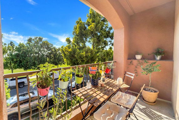 Balcony with potted plants and outdoor furniture overlooking a green area with trees and parked cars.