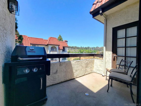 Balcony with barbecue grill and chairs, overlooking nearby buildings with red-tiled roofs.
