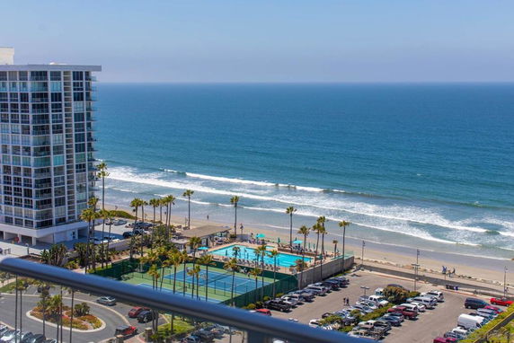 Panoramic view of a coastline with a high-rise building, tennis court, swimming pool, parking area, and ocean.