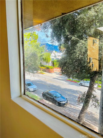 View from a window showing a street with parked cars, trees, and a distant house with mountains in the background.