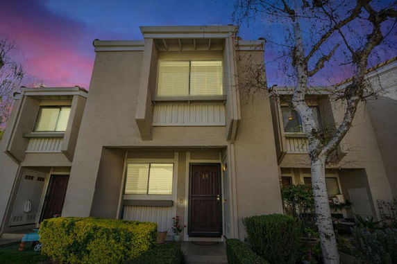 Front view of a two-story townhouse with a brown door and windows, surrounded by plants.