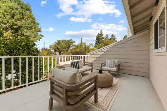 Balcony view showing a residential outdoor space with seating, a small table, and a railing overlooking trees and a clear sky.