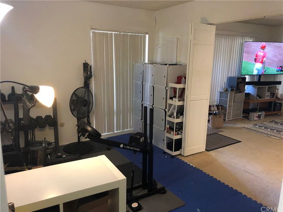 Room with exercise equipment including weights and a workout bench with a television and shelves in the background.