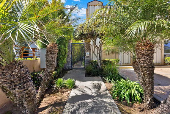Front view of a house with a pathway leading to a gate, surrounded by palm trees and greenery.