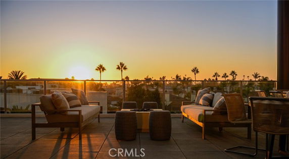 Outdoor seating area with couches and tables overlooking a sunset view with distant trees and buildings.